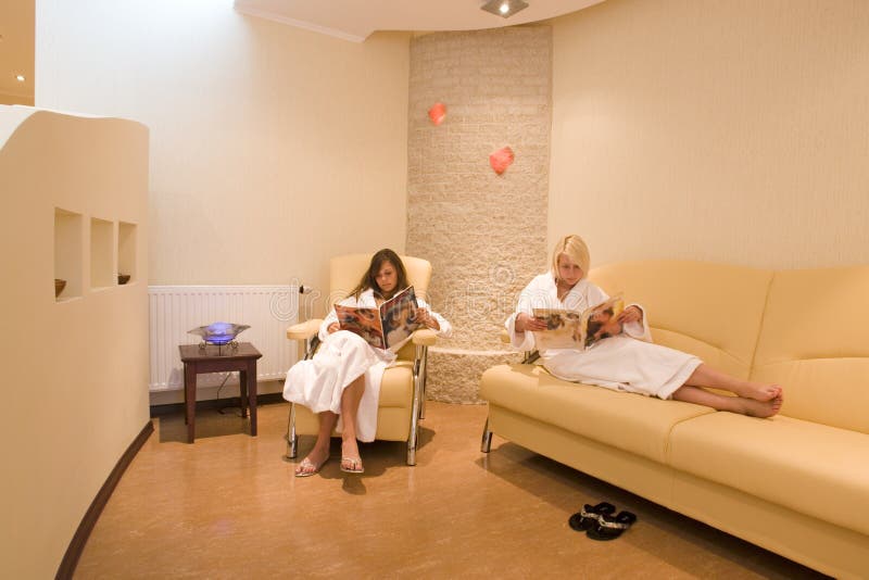 Two young women, dressed in white robes, reading while waiting in a lounge or waiting room at a resort spa. Two young women, dressed in white robes, reading while waiting in a lounge or waiting room at a resort spa.