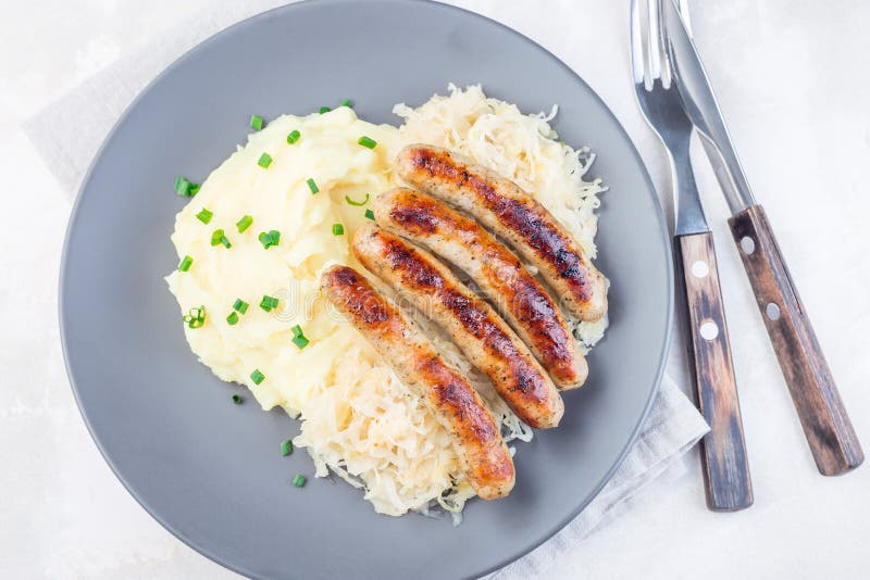 Rostad nurebergkorv serverad med surkål och krossad potatis, på grå platta, vågrät, ovanifrån royaltyfri bild Rostad nurebergkorv serverad med surkål och krossad potatis, på grå platta, vågrät, ovanifrån royaltyfri bild