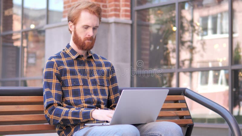 Rothaarige-Bart-Junge-Mann, der an Laptop, Sitzen im Freien auf Bank arbeitet