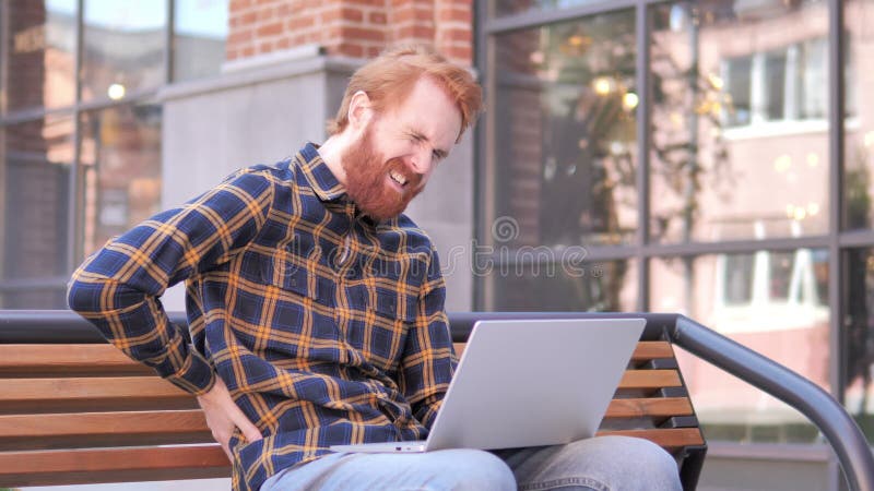 Rothaarige-Bart-junger Mann mit den Rückenschmerzen, die an dem Laptop im Freien arbeiten