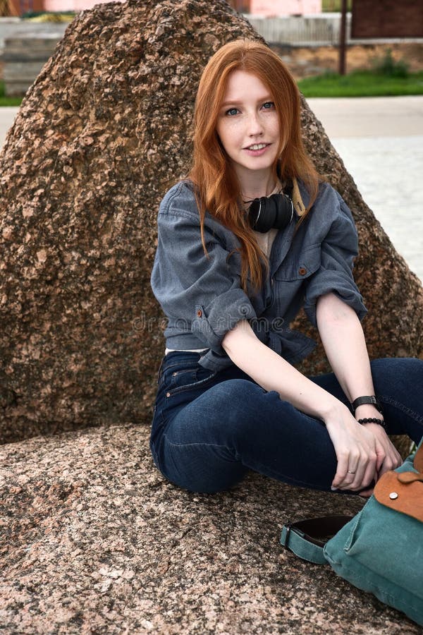 Rotkopfmädchen mit Freckles und blauen Augen in einem Denim-Hemd mit Kopfhörer am Nacken sitzen auf einem Stein und blickt in das stockfotografie