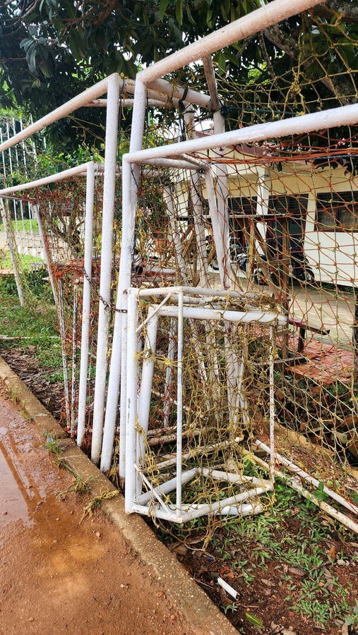 A row of weathered football goalposts with metal frames and rusted nets leans against a tree-lined edge. The ground is a mix of dirt and sparse grass. Behind the goals, a building with white walls and windows is visible, partially obscured by green foliage. The scene suggests an informal sports area, with equipment stored or set aside temporarily on a rustic path. A red motorcycle and wooden items are also seen near the building, indicating a casual, mixed-use space. Sparse use stock images, royalty-free photos and pictures