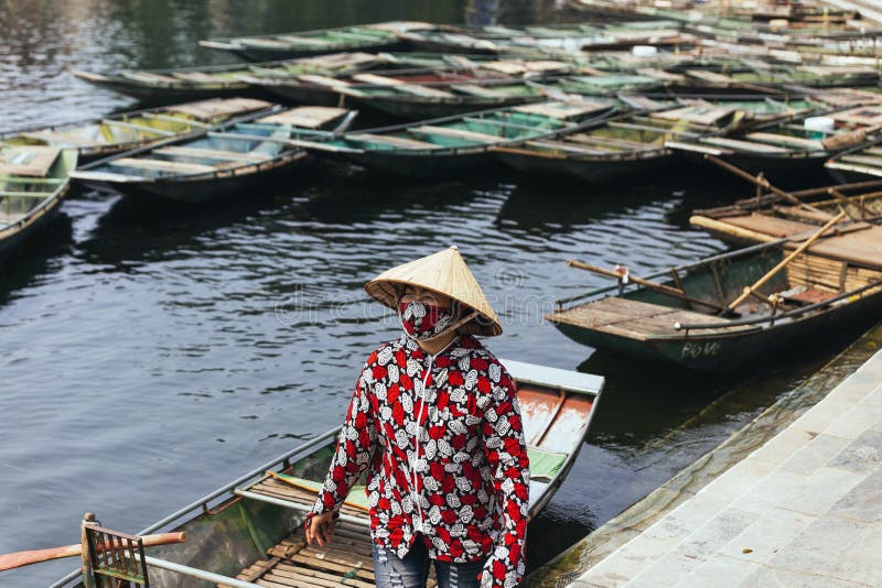 Rowing boat woman wearing red and white colors shirt, conical hat and mouth mask standing with empty boats over the river. royalty free stock photography Rowing boat woman wearing red and white colors shirt, conical hat and mouth mask standing with empty boats over the river. royalty free stock photography