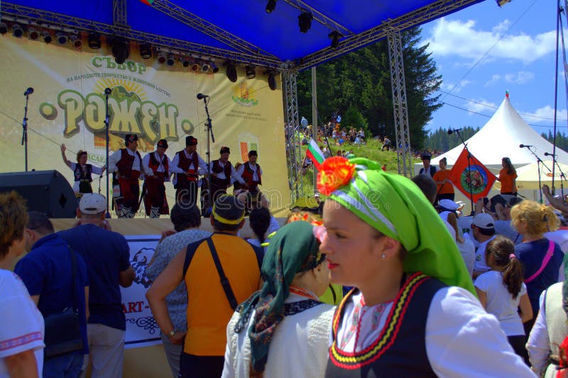 Spectators and women folklore group dressed in traditional authentic costumes in front of scene of Folklore Festival Rozhen in beautiful scenery of Rhodope Mountains.Every 4th year in July, Bulgarians of all over the country gather below the peak of Rozhen for a festival, where singers and instrumentalists sing and play on a several scenes. Spectators and women folklore group dressed in traditional authentic costumes in front of scene of Folklore Festival Rozhen in beautiful scenery of Rhodope Mountains.Every 4th year in July, Bulgarians of all over the country gather below the peak of Rozhen for a festival, where singers and instrumentalists sing and play on a several scenes.