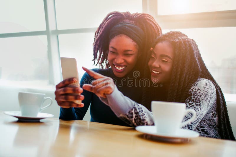 Cheerful young african women sit together. They smile and point on white phone. Models sit ar bringht window. Cheerful young african women sit together. They smile and point on white phone. Models sit ar bringht window