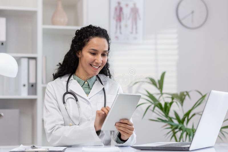 Professional Hispanic woman doctor sitting in a clinic office, smiling as she reviews patient data on a digital tablet. Modern healthcare setting. Professional Hispanic woman doctor sitting in a clinic office, smiling as she reviews patient data on a digital tablet. Modern healthcare setting.