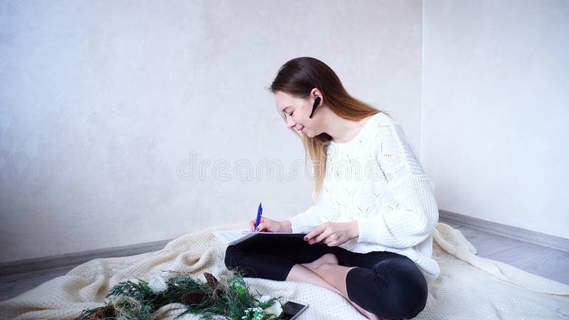 Organized women`s holiday organizer communicates with client on bluetooth headset and writes pen in folder with papers ideas and information about event. girl with good mood smiles and laughs, sits on floor on knitted plaid near Christmas pine wreath and flickering garland in bright room in daytime. European-looking Woman with long blond hair is dressed in white sweater and black pants. Concept of gadgets and modern technology, consumption and training of new devices, remote work and entrepreneurship, festive mood and New Year or Christmas Eve, happy and beautiful people, holiday attributes and symbols.r. Organized women`s holiday organizer communicates with client on bluetooth headset and writes pen in folder with papers ideas and information about event. girl with good mood smiles and laughs, sits on floor on knitted plaid near Christmas pine wreath and flickering garland in bright room in daytime. European-looking Woman with long blond hair is dressed in white sweater and black pants. Concept of gadgets and modern technology, consumption and training of new devices, remote work and entrepreneurship, festive mood and New Year or Christmas Eve, happy and beautiful people, holiday attributes and symbols.r