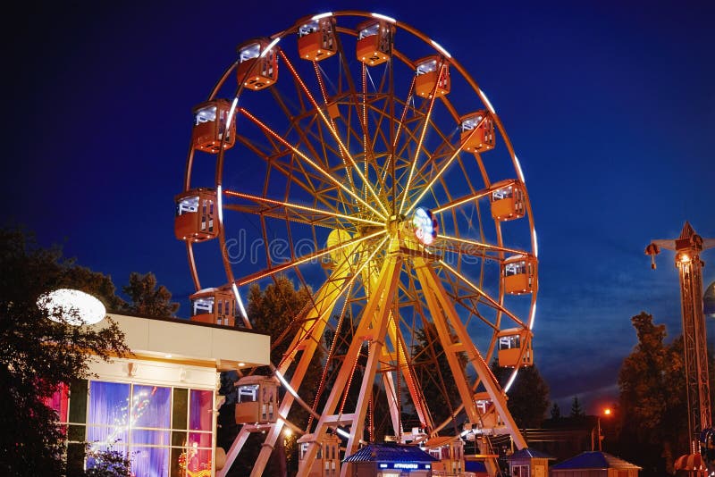 Rueda de hierro iluminada en parque de atracciones en una ciudad nocturna. Rueda de ferris iluminada en parque de atracciones en ciudad nocturna, carrusel, luz, carnaval, ocio, viajes, velada, diversión, paseo, movimiento, exterior, feria, exposición, turismo, vacaciones, celebración, alegría, atracción, redonda, familiar, tradicional, luminiscencia, círculo, luminosidad, verano, entretenimiento, luminosidad, luminosidad, luminancia, electricidad, brillo, color naranja, color amarillo, amarillo, luces, lunaparca, circular, circular. festival, riesgo