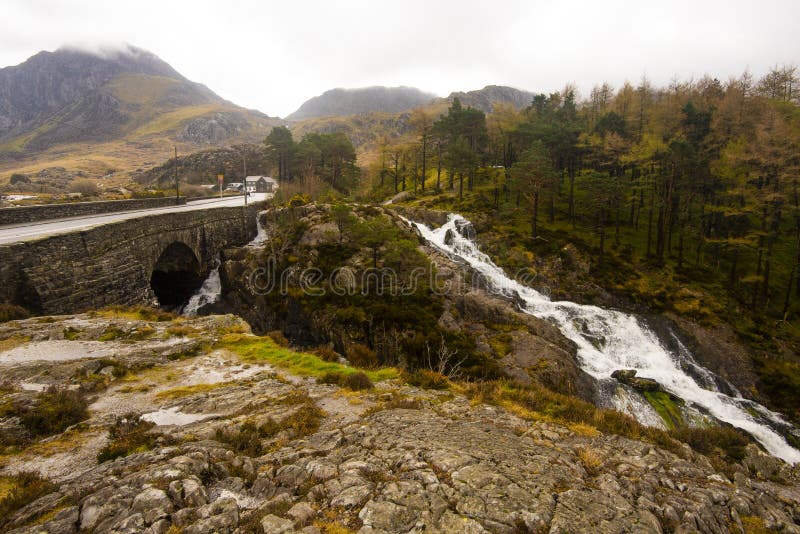 Rugged Welsh landscape, with a road and bridge over fast flowing river, rugged rocks, fir trees and low cloud ov er the mountains