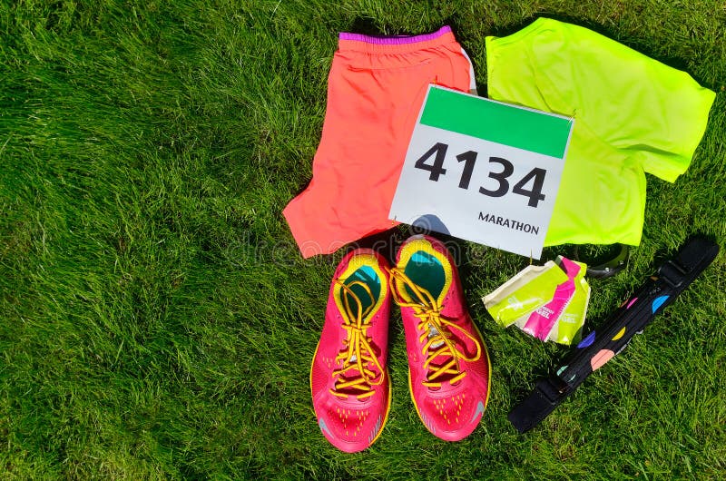Running shoes, marathon race bib (number), runners gear and energy gels on grass background