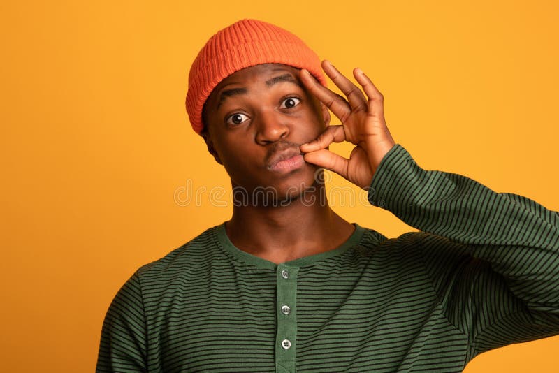 Keep Quiet. Young Black Hipster Guy In Orange Hat Making Zipping Mouth Gesture, Holding Fingers Near Lips, Funny African American Man Promising To Hold Secret, Standing On Yellow Background, Closeup. Keep Quiet. Young Black Hipster Guy In Orange Hat Making Zipping Mouth Gesture, Holding Fingers Near Lips, Funny African American Man Promising To Hold Secret, Standing On Yellow Background, Closeup