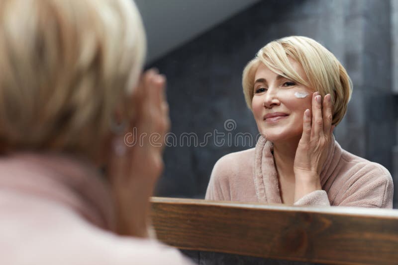 Rutina de cuidado de la piel. mujer madura utiliza crema cosmética en la piel de la cara delante del reflejo del espejo. imagen de archivo Rutina de cuidado de la piel. mujer madura utiliza crema cosmética en la piel de la cara delante del reflejo del espejo. imagen de archivo