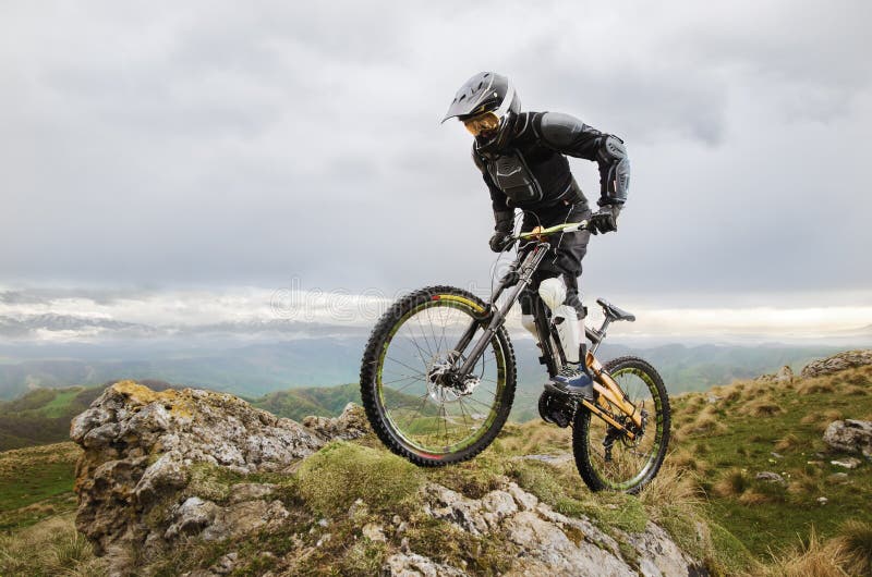 Ryder in full protective equipment on the mtb bike climbs on a rock against the backdrop of a mountain range and low