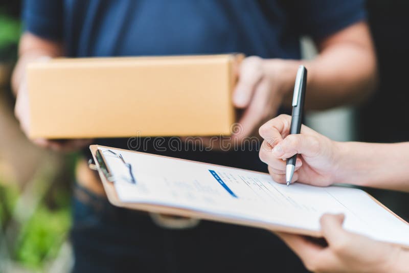 Close up shot of woman hand putting signature in clipboard to receive package from delivery man in background. Close up shot of woman hand putting signature in clipboard to receive package from delivery man in background