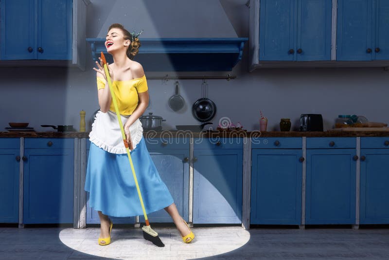 Retro pin up girl woman female housewife wearing yellow top, blue skirt and white apron holding mop singing and cleaning floor in stage light kitchen with blue cabinets and utensils. Housework concept. Retro pin up girl woman female housewife wearing yellow top, blue skirt and white apron holding mop singing and cleaning floor in stage light kitchen with blue cabinets and utensils. Housework concept