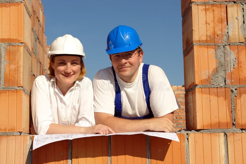 Couple wearing hard hats posing in their future house window opening resting on building plan. Couple wearing hard hats posing in their future house window opening resting on building plan