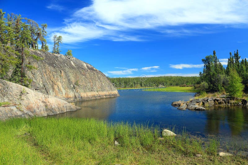 Just above Cameron Falls, the clear waters of the Cameron River flow slowly through a wide valley rimmed with reeds and forests - and the rounded outcroppings of structured Precambrian rock from the Canadian Shield, Hidden Lake Territorial Park, Northwest Territories, Canada. Just above Cameron Falls, the clear waters of the Cameron River flow slowly through a wide valley rimmed with reeds and forests - and the rounded outcroppings of structured Precambrian rock from the Canadian Shield, Hidden Lake Territorial Park, Northwest Territories, Canada.