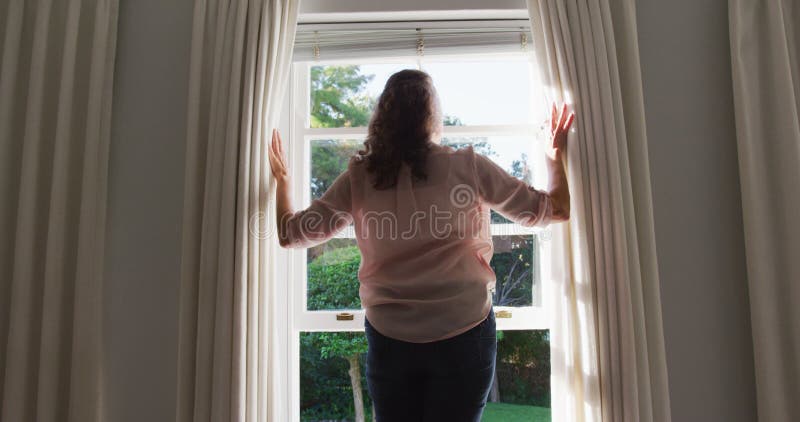 Rückansicht von Senior kaukasische Frau, die Vorhänge im sonnigen Raum zieht und aus dem Fenster schaut