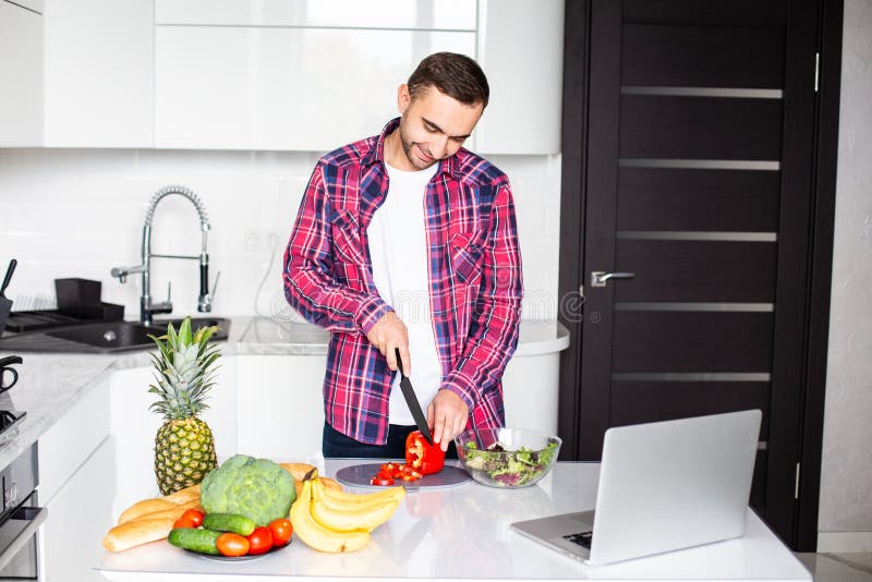 It`s so delicious. Casual happy young man preparing salad at home in loft kitchen and smiling, using laptop stock image