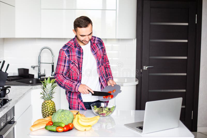 It`s so delicious. Casual happy young man preparing salad at home in loft kitchen and smiling, using laptop royalty free stock image