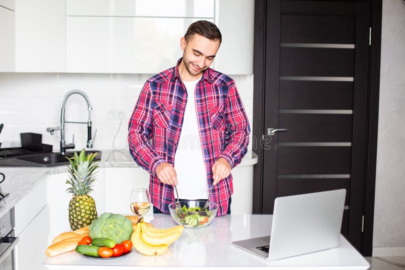 It`s so delicious. Casual happy young man preparing salad at home in loft kitchen and smiling, using laptop stock photography