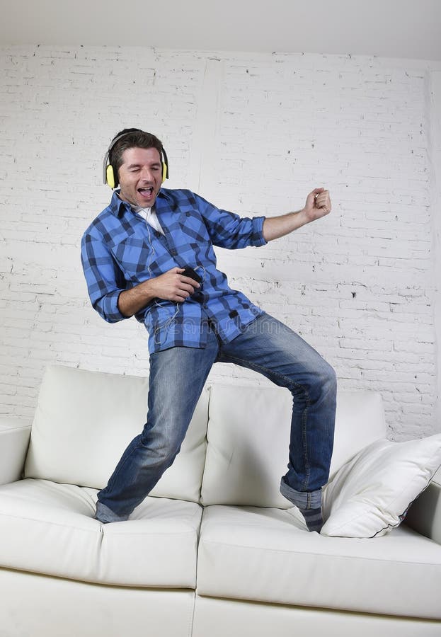 20s or 30s man jumped on couch listening to music on mobile phone with headphones playing air guitar