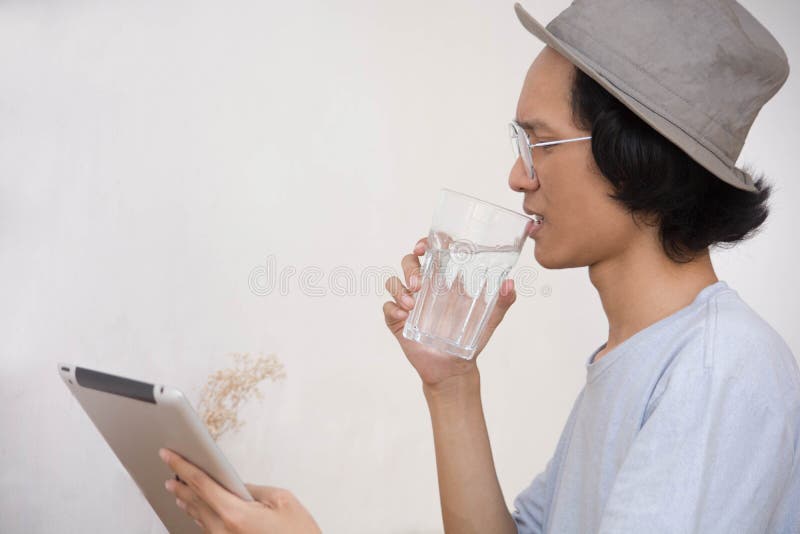 Young asian man with hat and glasses surprised at tablet or laptop while drink a glass of water stock photography Young asian man with hat and glasses surprised at tablet or laptop while drink a glass of water stock photography