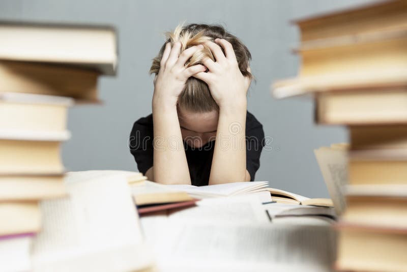 A sad boy of 9-10 years old in stress sits at a table with books and shares his hands behind his head. Gray background. Exams and