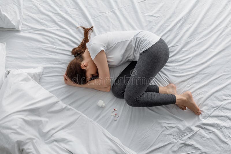 Sad despair young caucasian woman suffering from abdominal pain lying on bed with jar and scattered pills