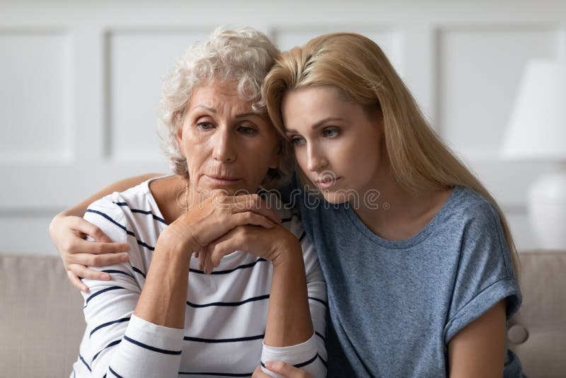 Sad grownup daughter sitting on sofa hugging desperate elderly mother stock photo