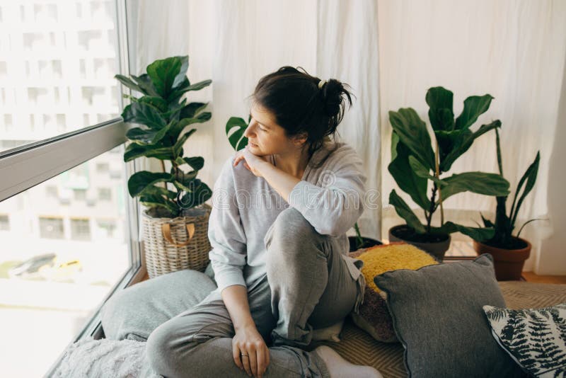 Sad hipster girl sitting at home during quarantine and looking at window. Stay home stay safe. Isolation at home to prevent virus