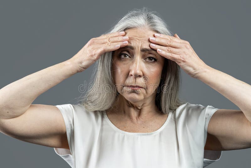 Sad old european lady with gray hair touches her face, wrinkles with hands, isolated on gray background