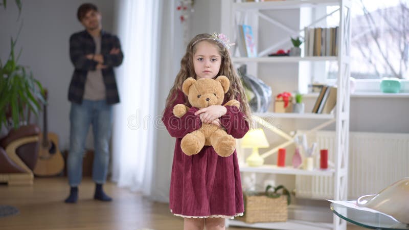 Sad pretty girl looking back at blurred man with crossed hands and looking at camera hugging teddy bear. Portrait of