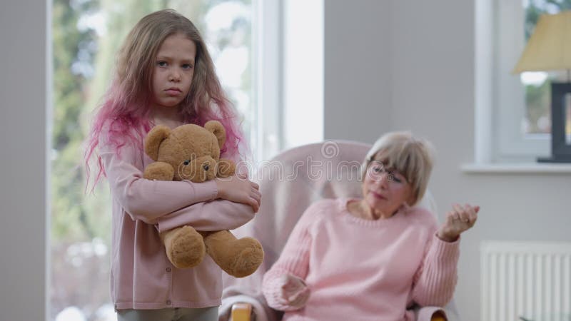 Sad pretty girl standing on the left hugging teddy bear looking at camera as dissatisfied senior woman scolding sitting