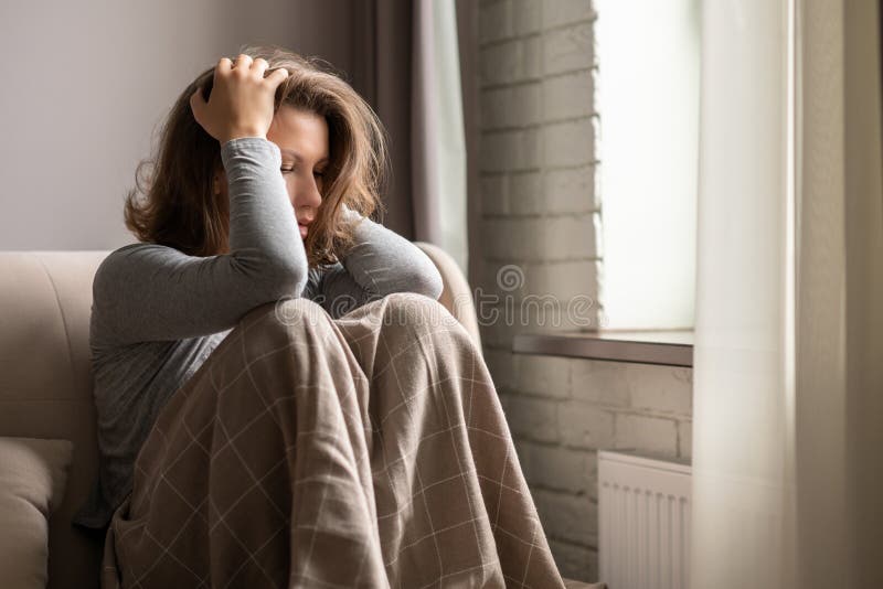 Sad scared woman in panic attack grabbing her head by hand. Lonely girl wrapped in a plaid sitting near window of apartments.