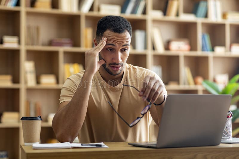 Sad tired arab male freelancer working on laptop and touching head, holding glasses, looking at computer screen royalty free stock photo