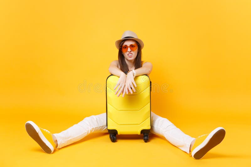 Sad traveler tourist woman in summer casual clothes, hat sit near suitcase on yellow orange background. Female