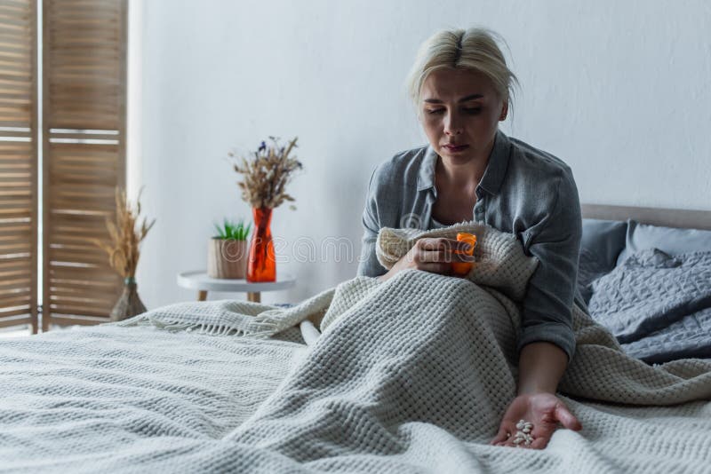 sad woman with climax looking at bottle with painkillers while sitting in bed under blanket,stock image