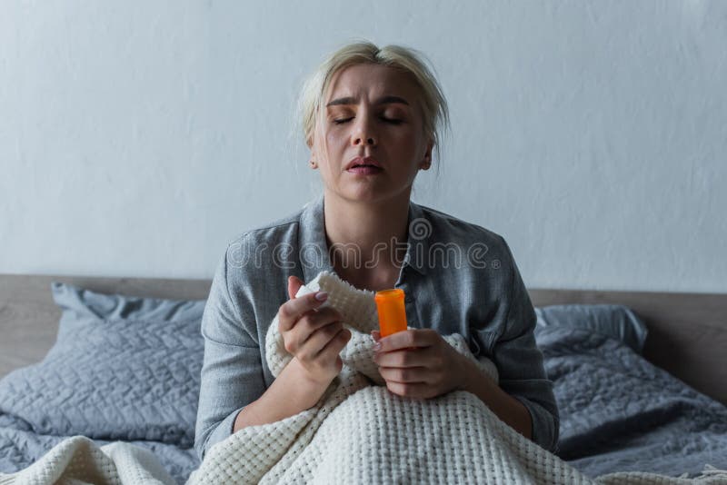 sad woman with climax sitting in bed and holding bottle with painkillers,stock image