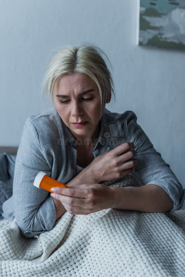 sad woman with climax sitting in bed while holding glass of water and painkillers,stock image