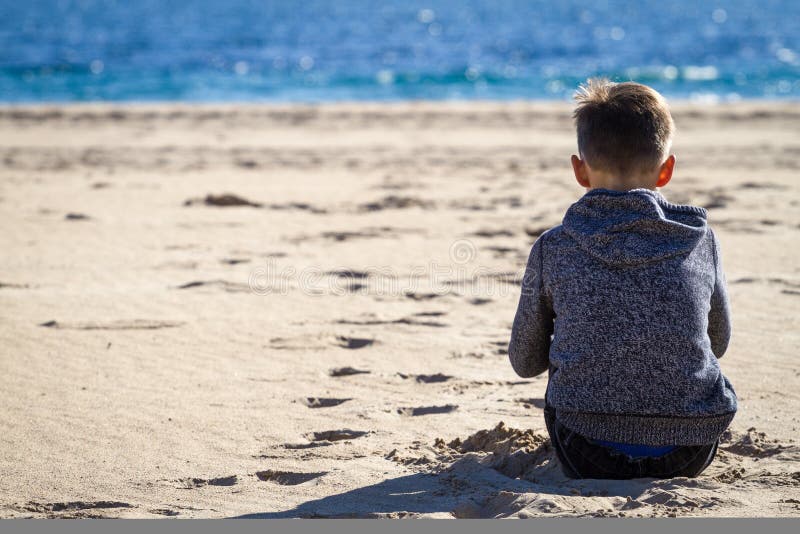 Sad young boy sitting on the beach, looking at sea and thinking