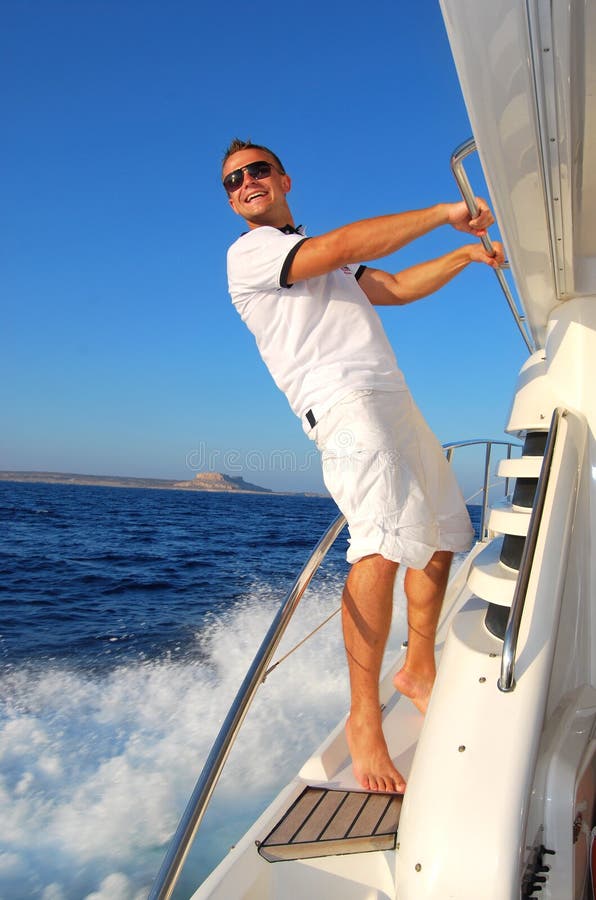 Young Sailor relaxing happily on the vacation sailboat yacht standing on a deck having a rest on summer boat over blue ocean wave splashes background. Young Sailor relaxing happily on the vacation sailboat yacht standing on a deck having a rest on summer boat over blue ocean wave splashes background