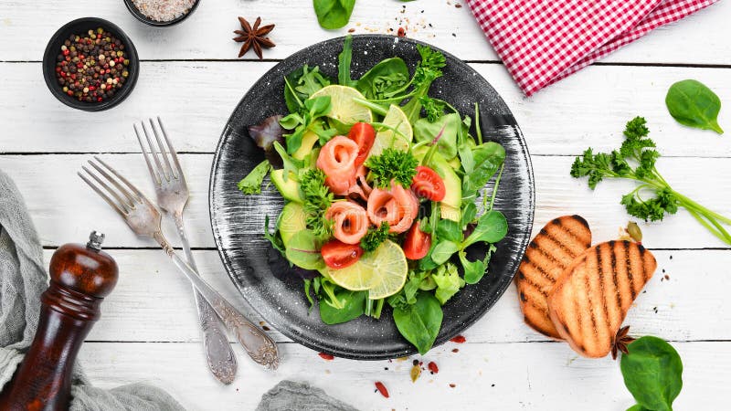 Salad with salmon, avocados, tomatoes and spinach. In a black plate on a wooden background Top view.