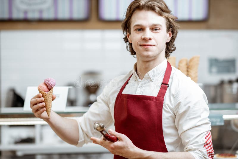 Portrait of a young and happy salesman in red apron standing with ice cream cone in the modern pastry shop. Portrait of a young and happy salesman in red apron standing with ice cream cone in the modern pastry shop