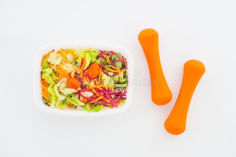 Sallad för den nya grönsaken i lunchask med orange hantlar övar utrustning på vit Aktiva sunda livsstilar, goda hälsor