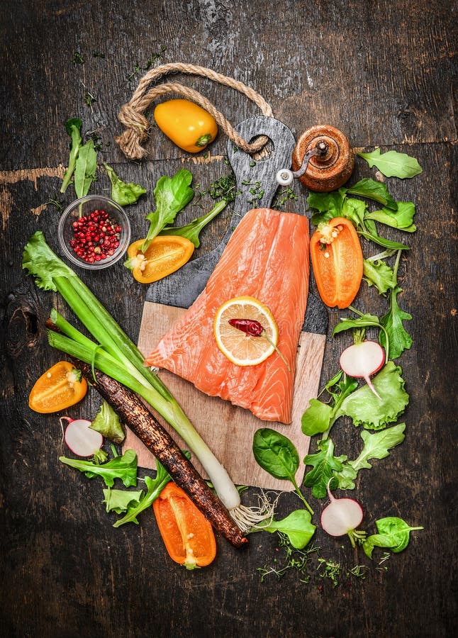 Salmon fish fillets on cutting board with fresh vegetables and spices ingredients on rustic wooden background, top view.