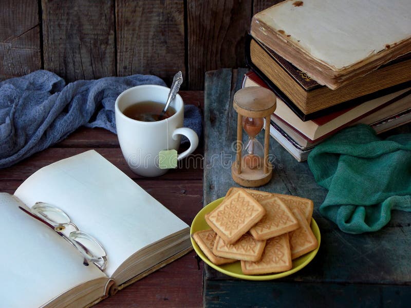 Sammansättningen av en bunt av gamla böcker, öppnar boken, tekoppar, exponeringsglas och plattor av sockerkakor på en träbakgrund