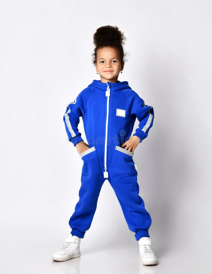 Full-growth portrait of self-confident dark-skinned afro-russian kid girl in blue trendy jumpsuit with pockets and zipper and white sneakers stands with her feet spread and hands in pockets. Full-growth portrait of self-confident dark-skinned afro-russian kid girl in blue trendy jumpsuit with pockets and zipper and white sneakers stands with her feet spread and hands in pockets