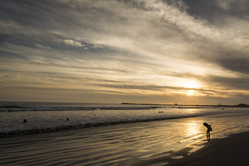 This wide, horizontal shot captures a serene sunset scene over the Pacific Ocean, likely in Riviera Nayarit, Mexico. The sun is low on the horizon, casting a golden glow across the water and reflecting on the wet, sandy beach. Gentle waves roll onto the shore, leaving foamy white lines. In the foreground, two silhouetted figures, possibly an adult and a child, stand on the wet sand, their reflections visible. Other smaller silhouettes of people are scattered along the distant waterline, with some individuals wading in the ocean. The sky above is a mix of muted blues, greys, and oranges, with elongated clouds stretching across the frame, creating a dramatic and peaceful atmosphere. A faint outline of land or a distant pier is visible on the far right horizon. Serene beach atmosphere stock images, royalty-free photos and pictures
