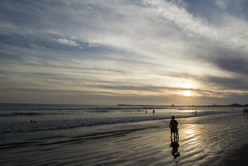 This wide, horizontal shot captures a serene sunset scene over the Pacific Ocean, likely in Riviera Nayarit, Mexico. The sun is low on the horizon, casting a golden glow across the water and reflecting on the wet, sandy beach. Gentle waves roll onto the shore, leaving foamy white lines. In the foreground, two silhouetted figures, possibly an adult and a child, stand on the wet sand, their reflections visible. Other smaller silhouettes of people are scattered along the distant waterline, with some individuals wading in the ocean. The sky above is a mix of muted blues, greys, and oranges, with elongated clouds stretching across the frame, creating a dramatic and peaceful atmosphere. A faint outline of land or a distant pier is visible on the far right horizon. Serene beach atmosphere stock images, royalty-free photos and pictures